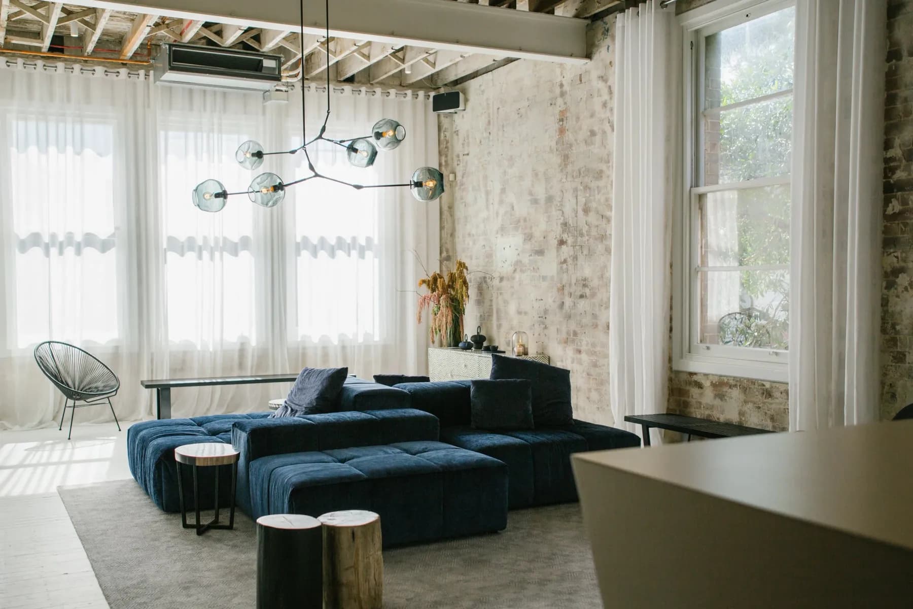A lounge with a navy sofa, glass pendant lights, sheer curtains and a whitewashed brick wall