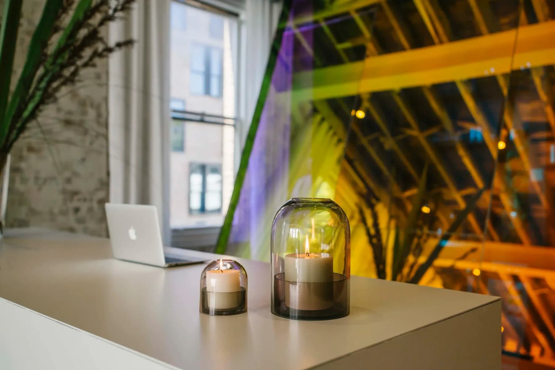 Two candles under glass domes in front of a window, with coloured light across the curtain